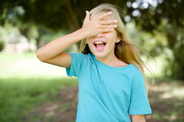 Caucasian little kid girl wearing blue T-shirt standing outdoors smiling and laughing with hand on face covering eyes for surprise. Blind concept.