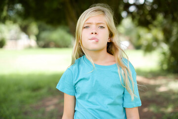 Caucasian little kid girl wearing blue T-shirt standing outdoors making grimace and crazy face, screaming out of control, funny lunatic expressing freedom and wild.