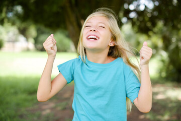 Caucasian little kid girl wearing blue T-shirt standing outdoors being excited for success with raised arms and closed eyes celebrating victory. Winner concept.