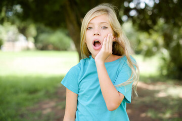 Shocked Caucasian little kid girl wearing blue T-shirt standing outdoors looks with great...