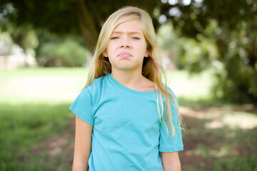 Displeased upset Caucasian little kid girl wearing blue T-shirt standing outdoors frowns face as going to cry, being discontent and unhappy as can't achieve goals,  Disappointed model has troubles