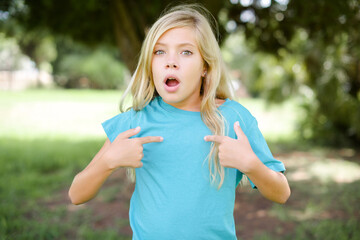 Embarrassed Caucasian little kid girl wearing blue T-shirt standing indicates at herself with puzzled expression, being shocked to be chosen to participate in competition, hesitates about something
