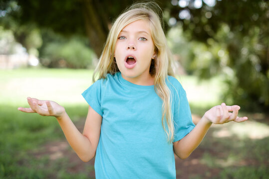 Frustrated Caucasian little kid girl wearing blue T-shirt standing outdoors feels puzzled and hesitant, shrugs shoulders in bewilderment, keeps mouth widely opened, doesn't know what to do.