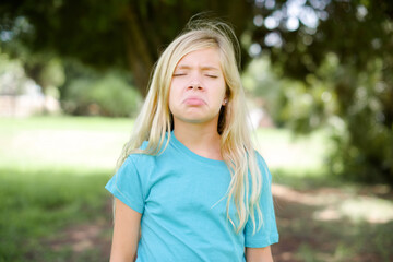 Dismal gloomy rejected Caucasian little kid girl wearing blue T-shirt standing outdoors has problems and difficulties, curves lower lip and closes eyes in despair, being in depression