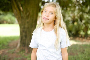 Obraz premium Funny Caucasian little kid girl wearing whiteT-shirt standing outdoors makes grimace and crosses eyes plays fool has fun alone sticks out tongue.