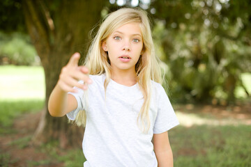 Shocked Caucasian little kid girl wearing whiteT-shirt standing outdoors points at you with stunned expression