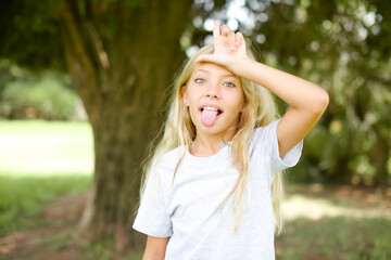 Caucasian little kid girl wearing whiteT-shirt standing outdoors gestures with finger on forehead makes loser gesture makes fun of people shows tongue