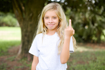 Caucasian little kid girl wearing whiteT-shirt standing outdoors shows middle finger bad sign asks not to bother. Provocation and rude attitude. - obrazy, fototapety, plakaty