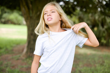 Caucasian little kid girl wearing whiteT-shirt standing outdoors stressed, anxious, tired and frustrated, pulling shirt neck, looking frustrated with problem