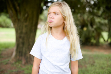 Dissatisfied Caucasian little kid girl wearing whiteT-shirt standing outdoors purses lips and has unhappy expression looks away stands offended. Depressed frustrated model.