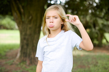 Caucasian little kid girl wearing whiteT-shirt standing outdoors purses lip and gestures with hand, shows something very little.