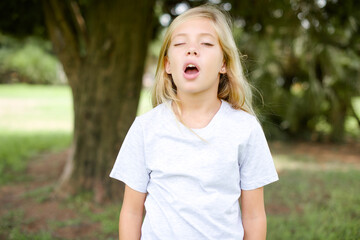 Caucasian little kid girl wearing whiteT-shirt standing outdoors yawns with opened mouth stands....