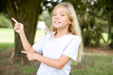 Caucasian little kid girl wearing whiteT-shirt standing outdoors points at copy space indicates for advertising gives right direction