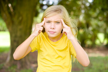 Serious concentrated beautiful Caucasian little kid girl wearing yellow T-shirt keeps fingers on temples, tries to ease tension, gather with thoughts and remember important information for exam