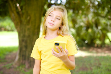 beautiful Caucasian little kid girl wearing yellow T-shirt standing outdoors holds mobile phone uses high speed internet and social networks has online communication. Modern technologies concept
