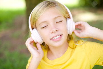 beautiful Caucasian little kid girl wearing yellow T-shirt standing outdoors with headphones on her...