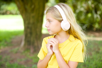 beautiful Caucasian little kid girl wearing yellow T-shirt standing outdoors wears stereo headphones listening to music concentrated and looking aside with interest.