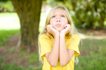Naklejka premium Portrait of sad beautiful Caucasian little kid girl wearing yellow T-shirt standing outdoors hands face look empty space