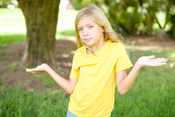 Naklejka na ściany i meble Careless attractive beautiful Caucasian little kid girl wearing yellow T-shirt standing outdoors shrugging shoulders, oops.