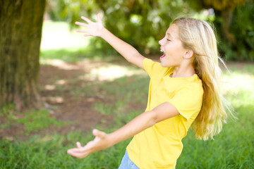 Obraz premium Funny astonished beautiful Caucasian little kid girl wearing yellow T-shirt standing outdoors look empty space with arms opened ready to catch something.