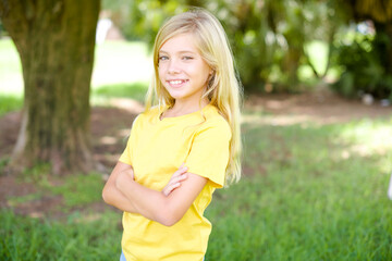 Fototapeta premium Portrait of beautiful Caucasian little kid girl wearing yellow T-shirt standing outdoors standing with folded arms and smiling