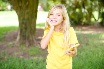 Fototapeta premium Afraid funny beautiful Caucasian little kid girl wearing yellow T-shirt standing outdoors holding telephone and bitting nails