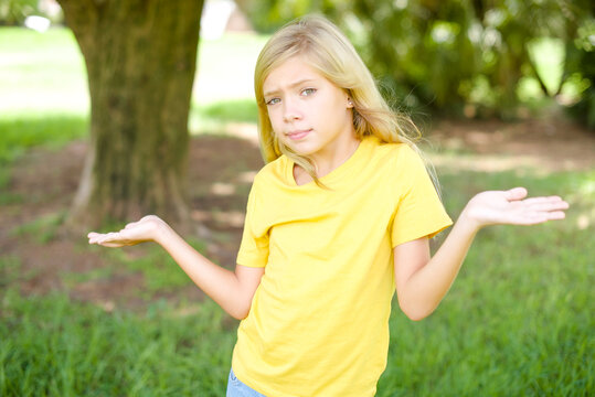 Careless attractive beautiful Caucasian little kid girl wearing yellow T-shirt standing outdoors shrugging shoulders, oops.