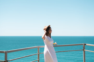 Young woman in white dress and hat stands by the sea.