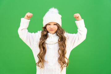 A girl in a warm white sweater and hat shows her fist and is very angry. A disgruntled baby with curly hair on an isolated background.