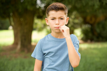 Fototapeta premium beautiful Caucasian little kid boy wearing blue T-shirt standing outdoors mouth and lips shut as zip with fingers. Secret and silent, taboo talking.