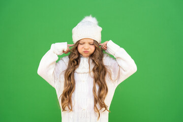 A little girl in a winter white sweater plugged her ears with her fingers and pouted her cheeks with displeasure. a child in winter clothes on an isolated background. A child with a cap and curly hair