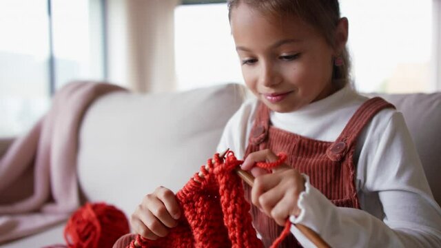 Little girl sitting on sofa and learning to knit indoors at home.