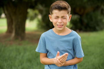 Caucasian little kid boy wearing blue T-shirt standing outdoor Suffering pain on hands and fingers, arthritis inflammation - obrazy, fototapety, plakaty