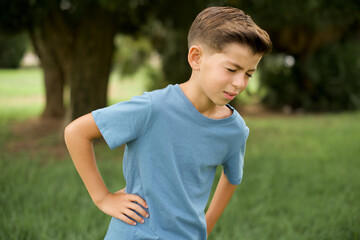 Caucasian little kid boy wearing blue T-shirt standing outdoor Suffering of backache, touching back...