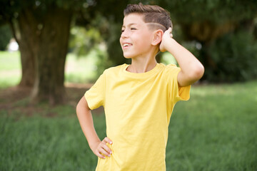 Caucasian little kid boy wearing yellow T-shirt standing outdoor  confuse and wonder about question. Uncertain with doubt, thinking with hand on head. Pensive concept.