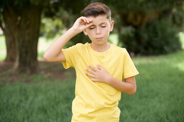 Caucasian little kid boy wearing yellow T-shirt standing outdoor  Touching forehead for illness and fever, flu and cold, virus sick.