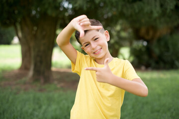 Naklejka premium Caucasian little kid boy wearing yellow T-shirt standing outdoor making finger frame with hands. Creativity and photography concept.
