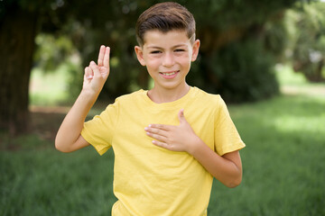 Caucasian little kid boy wearing yellow T-shirt standing outdoor  smiling swearing with hand on chest and fingers up, making a loyalty promise oath.