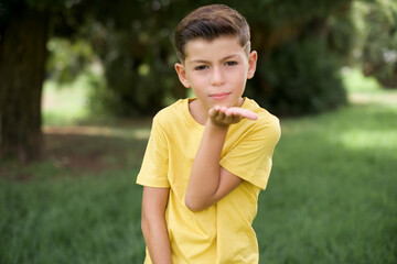 Fototapeta premium Caucasian little kid boy wearing yellow T-shirt standing outdoor looking at the camera blowing a kiss with hand on air being lovely and sexy. Love expression.