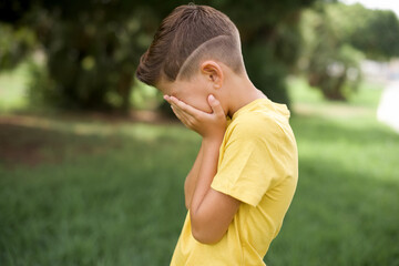 Sad  Caucasian little kid boy wearing yellow T-shirt standing outdoor  crying covering her face with her hands. © Roquillo