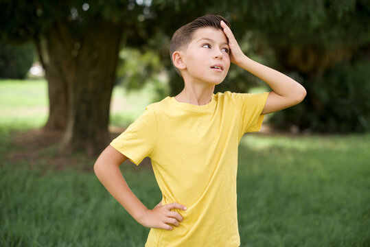 Caucasian little kid boy wearing yellow T-shirt standing outdoor  surprised with hand on head for mistake, remember error. Forgot, bad memory concept.
