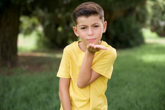 Caucasian little kid boy wearing yellow T-shirt standing outdoor  looking at the camera blowing a kiss with hand on air being lovely and sexy. Love expression.