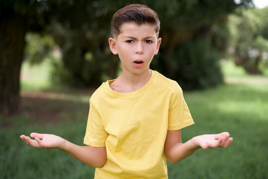 Frustrated Caucasian little kid boy wearing yellow T-shirt standing outdoor  feels puzzled and hesitant, shrugs shoulders in bewilderment, keeps mouth widely opened, doesn't know what to do.