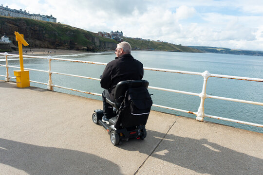 Disabled man using Mobility scooter to enable himself to independent and free to get out and about whilst on vacation sitting enjoying the sea breeze.