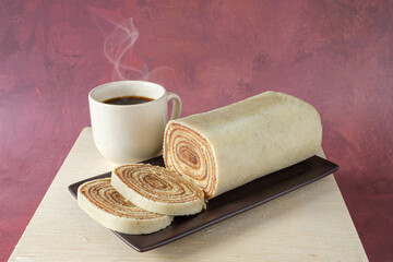 Sliced bolo de rolo on a brown plate, next to a cup of coffee.