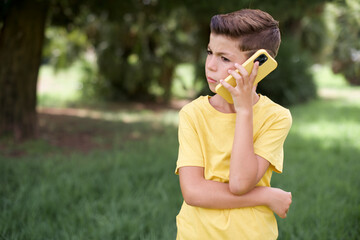 Sad Caucasian little kid boy wearing yellow T-shirt standing outdoors  talking on smartphone....