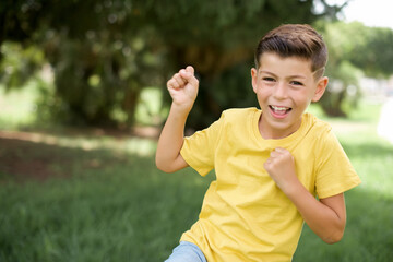 Profile photo of excited Caucasian little kid boy wearing yellow T-shirt standing outdoors raising...
