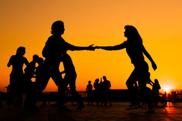 Man and woman couple silhouettes dancing against warm sunset orange sky on quay at evening. Group dance, romantic, love, summer outdoor activity and vacation concept