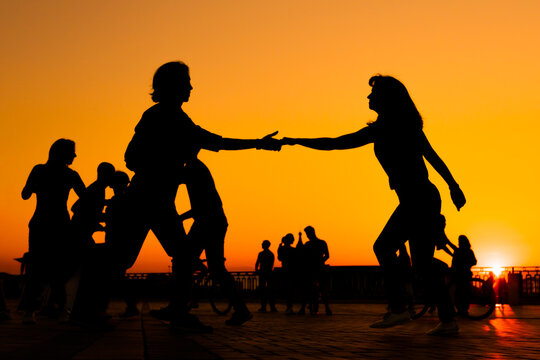 Man and woman couple silhouettes dancing against warm sunset orange sky on quay at evening. Group dance, romantic, love, summer outdoor activity and vacation concept