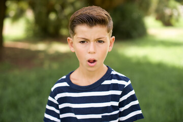 Shocked beautiful Caucasian little kid boy wearing stripped T-shirt standing outdoors stares bugged...